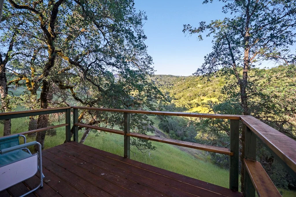 A wooden balcony or deck with a green metal chair, overlooking a lush green landscape with trees and rolling hills under a clear blue sky.