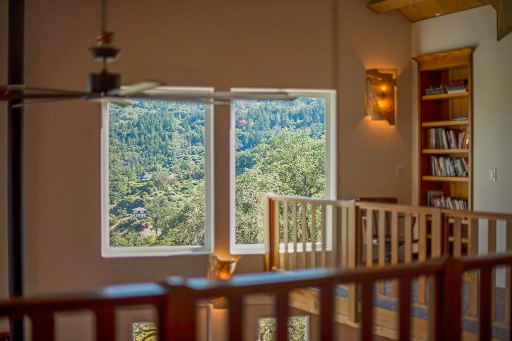 Interior of a room with a large window showing a view of a lush, green, mountainous landscape. There is a wooden bookshelf, a wall-mounted light, and a wooden railing inside the room.