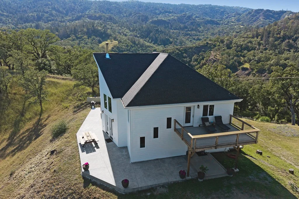 A white house with a black roof situated on a grassy hill, surrounded by trees and mountain scenery, with a wooden deck, outdoor furniture, and potted plants.