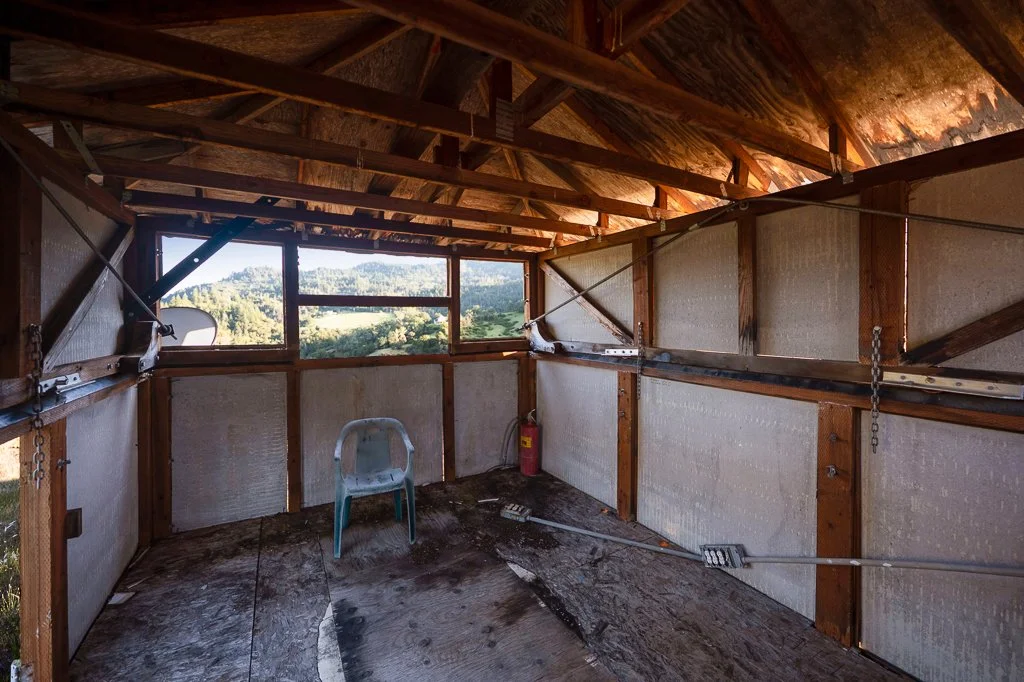 Empty unfinished wooden room with insulation, a plastic chair, a fire extinguisher, and a ladder, overlooking a scenic landscape through a large rectangular window.