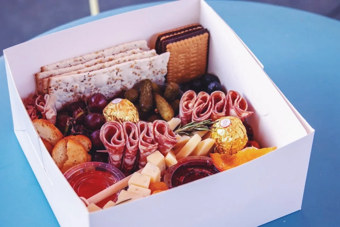 A white box filled with assorted snacks including crackers, cured meats, cheese cubes, grapes, blueberries, and chocolates.