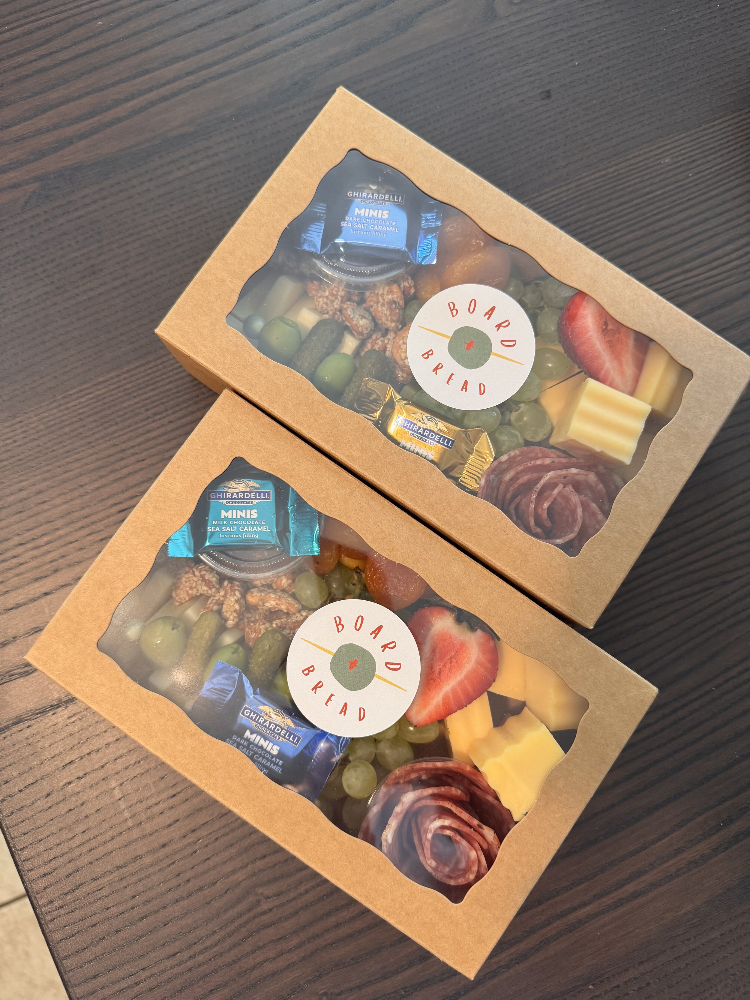 Two brown boxes of assorted cheeses, fruits, crackers, and snacks with a 'Board & Bread' label on top, placed on a dark wooden surface.