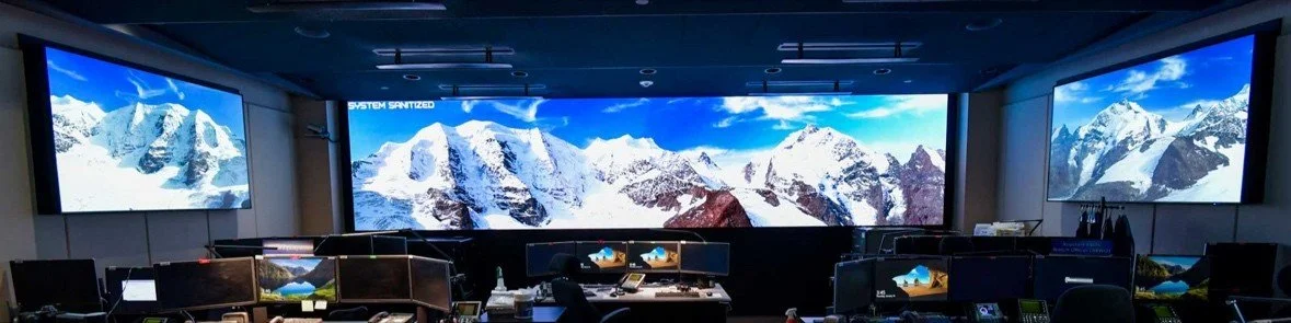A control room with multiple computer monitors and large screens showing snow-covered mountain peaks. This photo highlights one of Todd Haller's projects