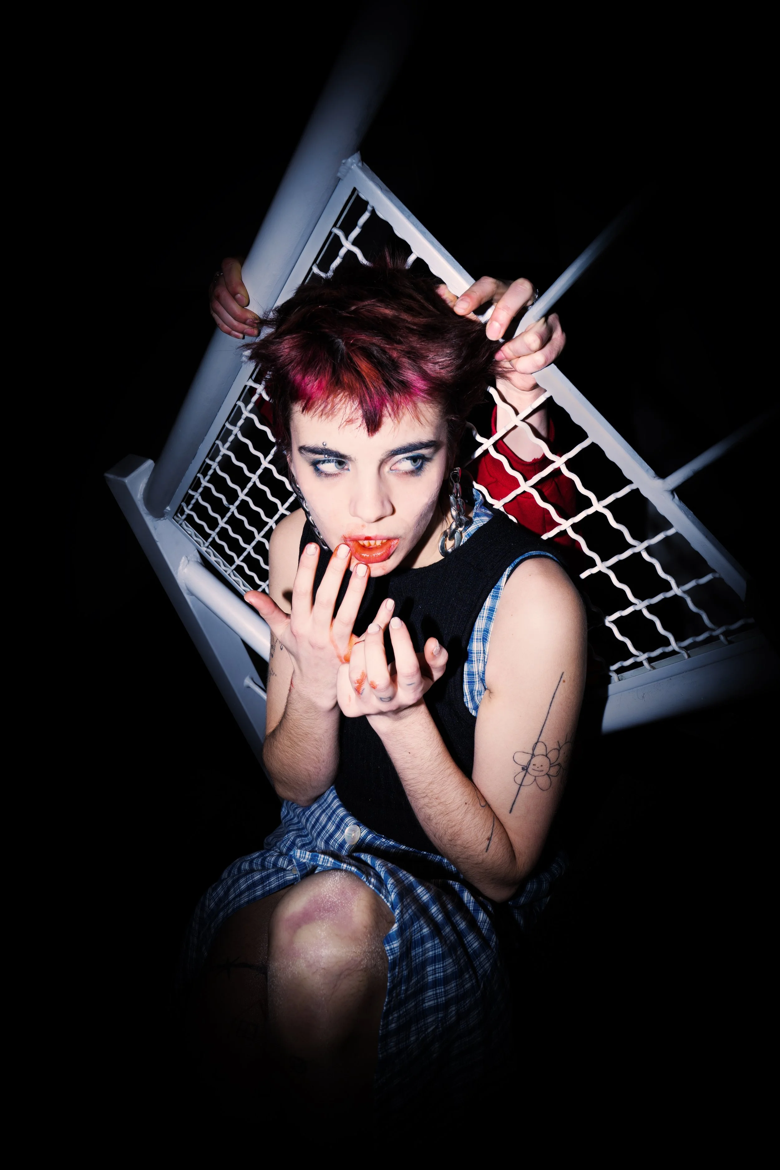 A young woman with colorful hair, makeup, and tattoos, sitting against a black background, gazing with an intense expression, with her hands near her face and a white metal fence behind her.