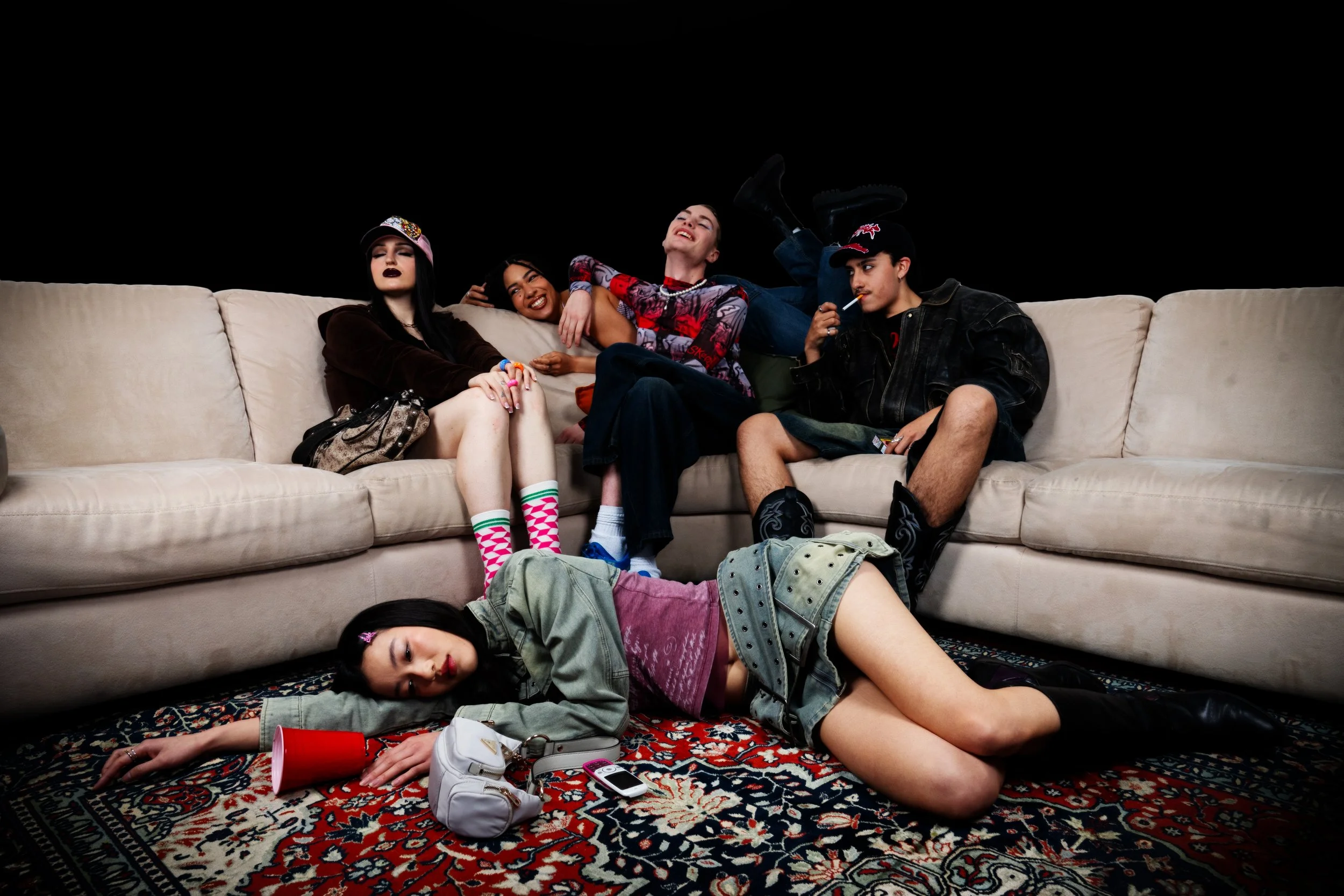 Group of five young women sitting on a beige couch, with one lying on the floor, in a room with black walls. They are dressed in casual clothing, some wearing hats and socks, and appear to be socializing and relaxing.