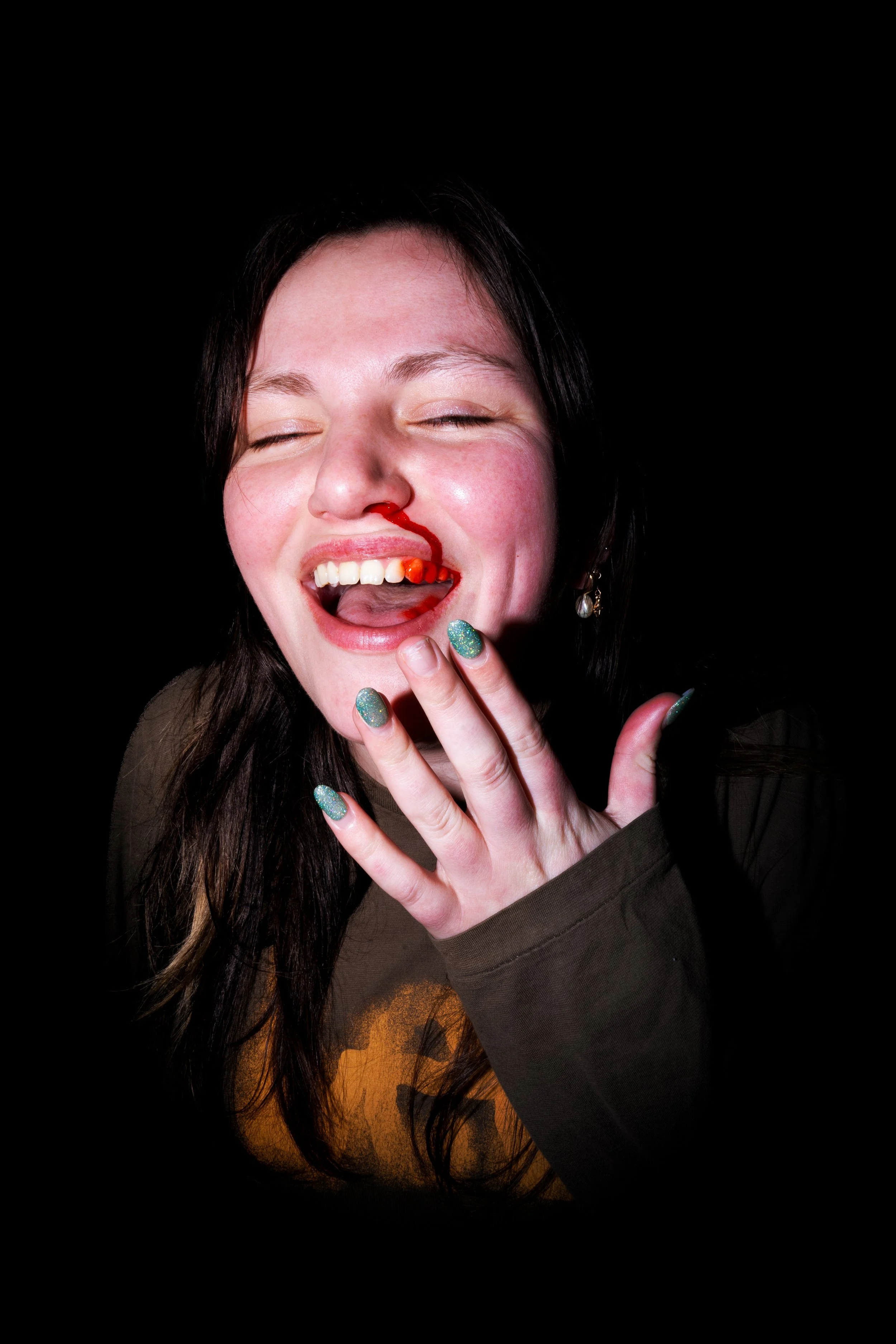 A woman with dark hair, smiling, with her eyes closed, has ketchup on her nose, holding her fingers near her mouth, with a playful expression, wearing a dark shirt and glittery nail polish.