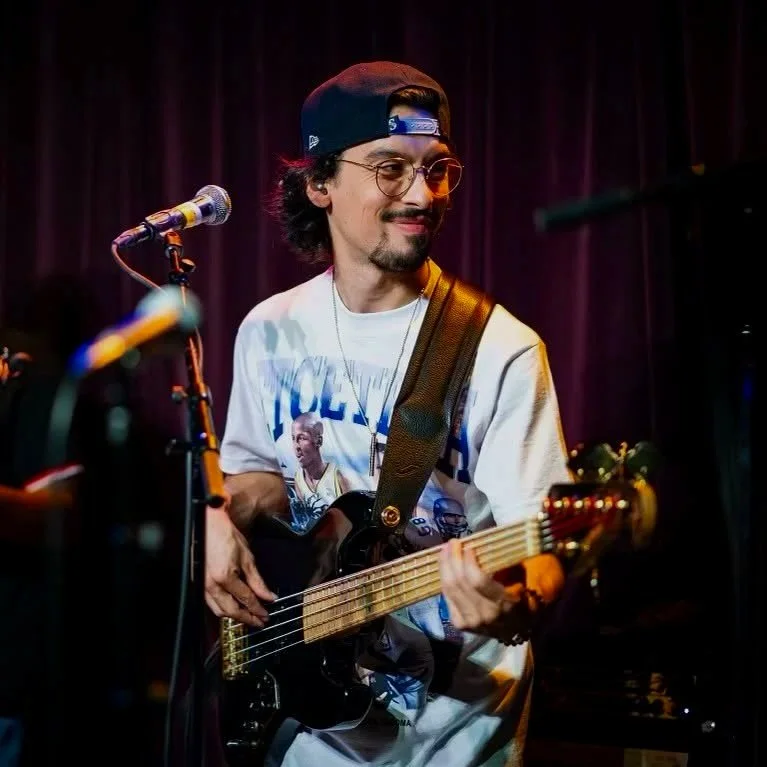 A man with glasses, a backwards baseball cap, and a goatee, playing an acoustic guitar on stage with a purple curtain background.