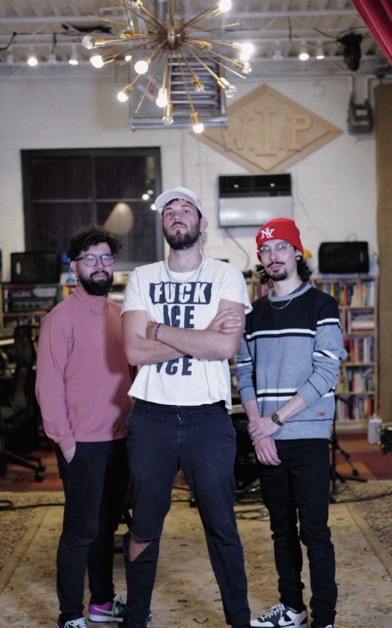 Three men standing together in a room with bookshelves, a window, and musical equipment. The man in the middle is wearing a white t-shirt with text, a white cap, and crosses his arms. The man on the left is wearing a pink shirt and glasses, and the m