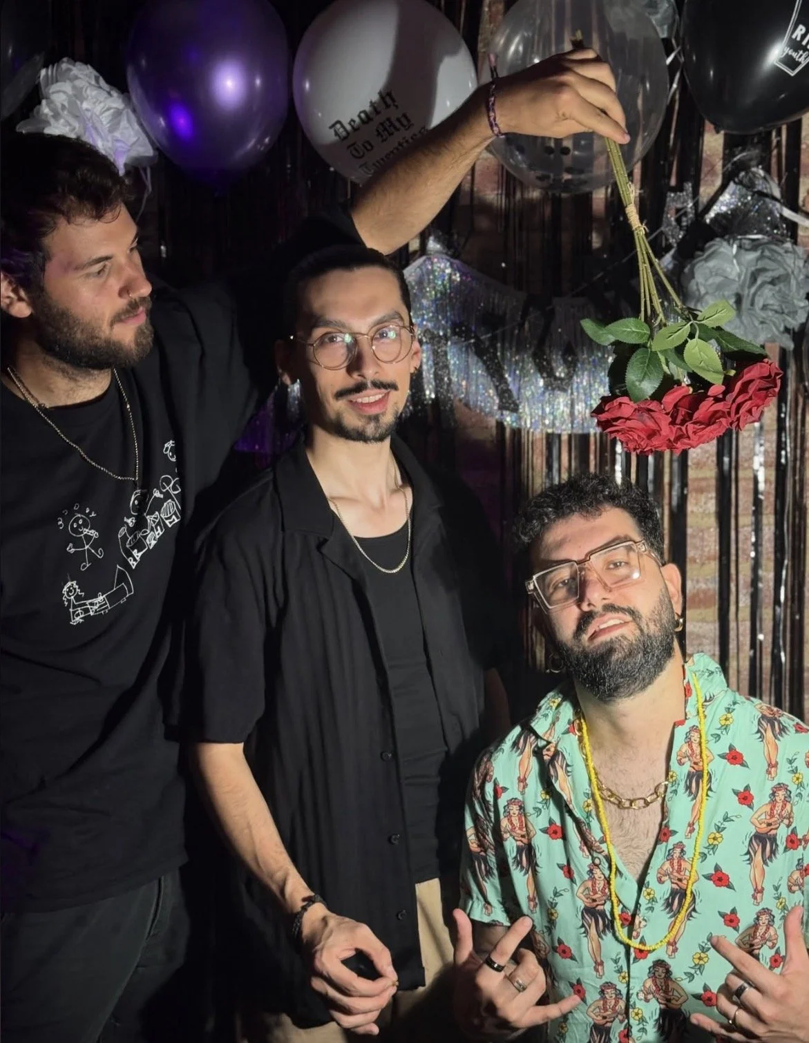 Three men at a party with black, purple, and white balloons, some with writing, and a hanging flower decoration. The room has a dark, festive background with shiny, metallic decorations.