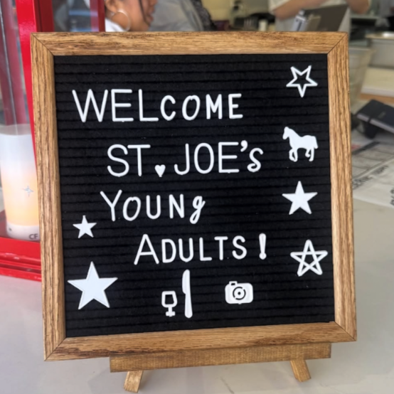 A black felt letter board with a wooden frame, featuring a welcoming message for young adults, surrounded by stars, a horse, and icons of a wine glass, a knife and fork, and a camera.