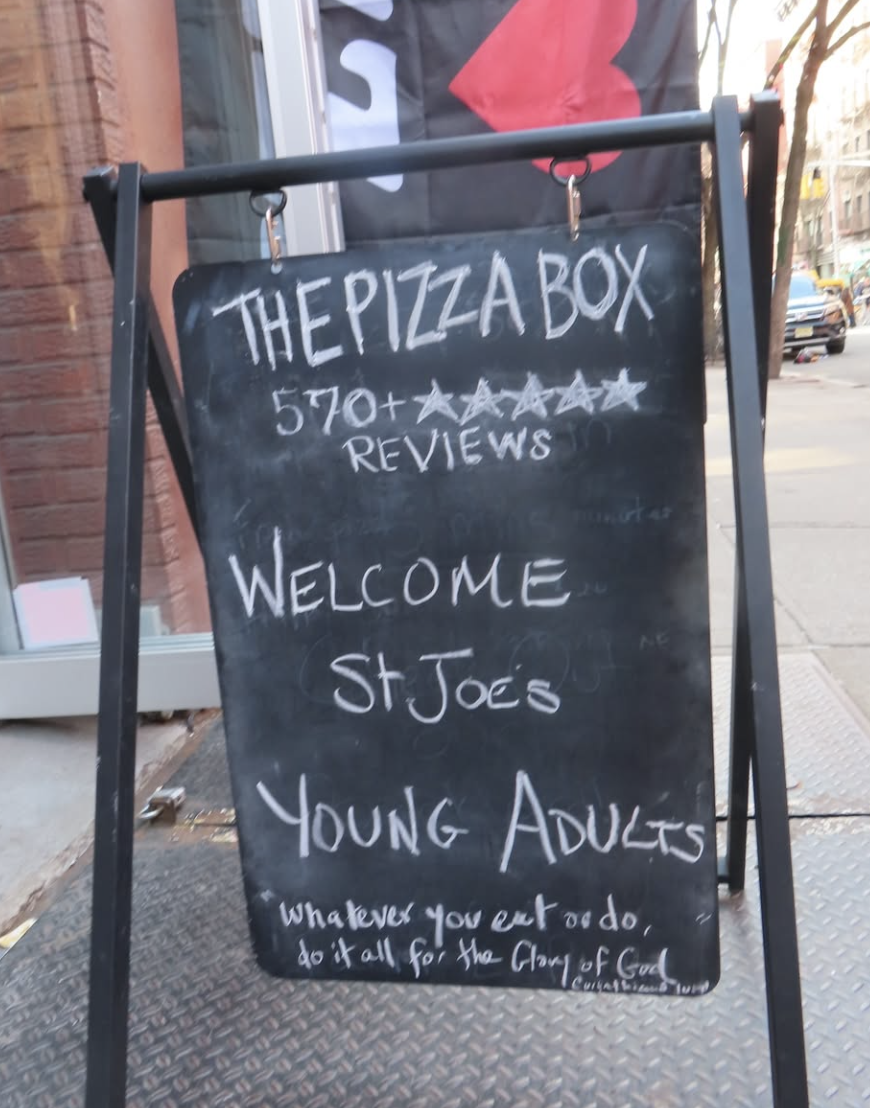 A black chalkboard sign outside a restaurant with white handwritten text that reads, "The Pizza Box, 570+ Reviews," and a welcoming message for St. Joe's Young Adults with a quote about doing everything for the glory of God at the bottom.
