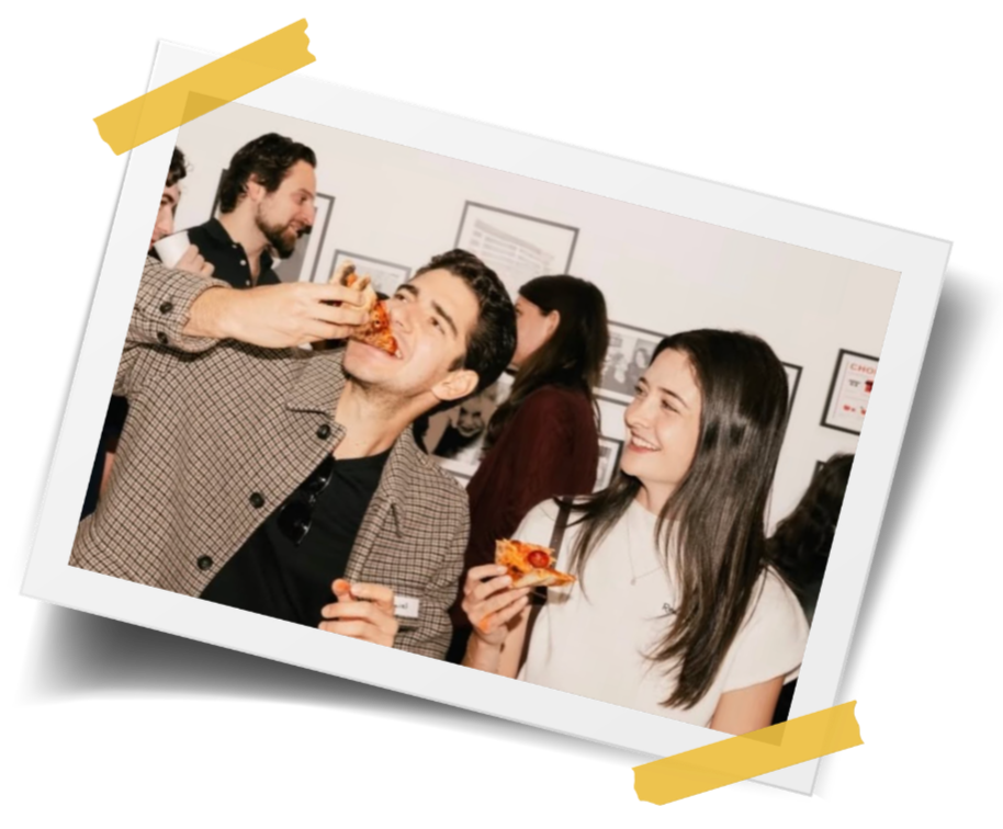 A group of people at a social gathering, with a man eating a slice of pizza while a woman next to him looks on and smiles, holding her own slice of pizza.