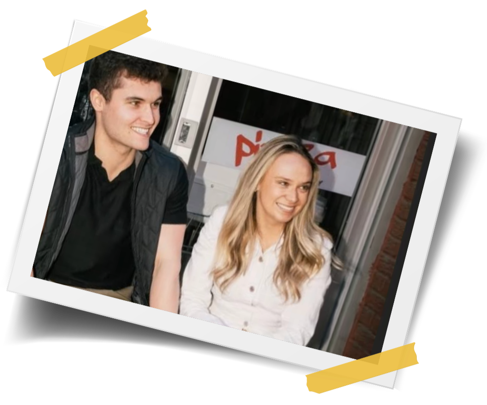 A young man and woman sitting together indoors, smiling and talking, with a pizza restaurant sign in the background.