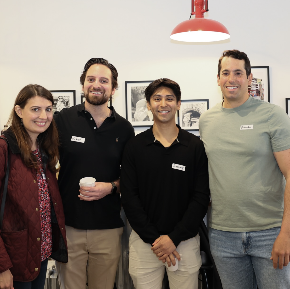 Four people standing together in an indoor setting, smiling at the camera, with artwork on the wall behind them and a red hanging light above.