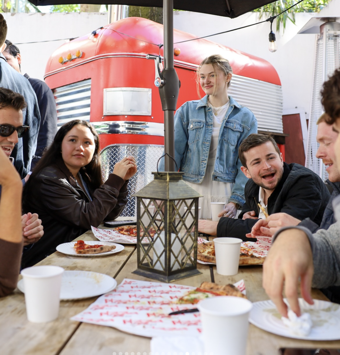 Group of young adults enjoying pizza outdoors under a canopy, with a red vintage camper in the background.