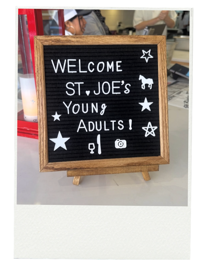A blackboard sign with a wooden frame displaying the message "Welcome St. Joe's Young Adults!" with decorative stars, a horse, a wine glass, a fork and knife, and a camera icons, on a white table in a restaurant or cafe.