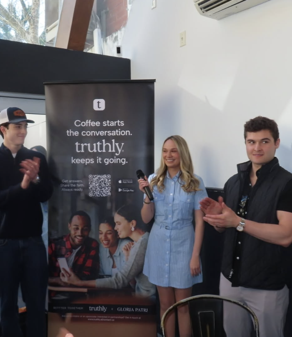 Three people are standing near a promotional banner at an indoor event. The woman in the middle is holding a microphone and smiling. The two men on either side are clapping. The banner advertises a faith-based app called 'truthly' with the slogan 'Coffee starts the conversation. truthly keeps it going.'