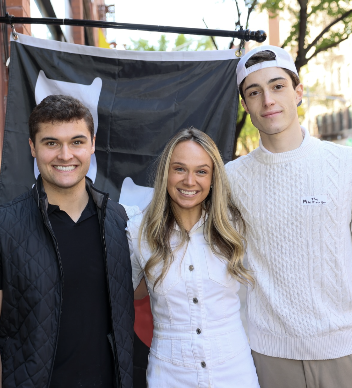 Three young adults smiling outdoors in front of a black flag with a white symbol, possibly at a rally or event, with trees and buildings in the background.