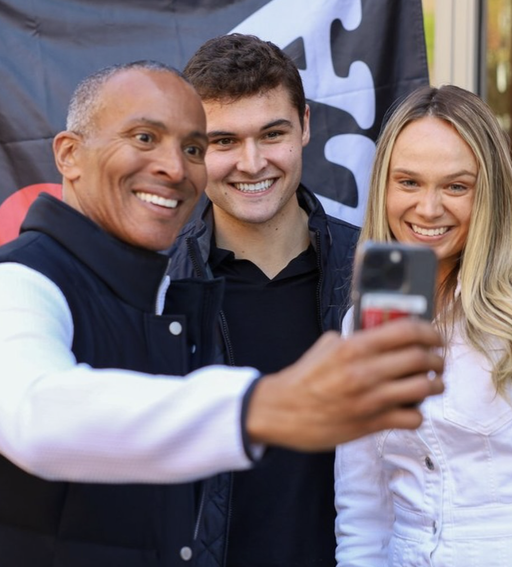 Three people smiling and taking a selfie outdoors, with a black banner in the background.