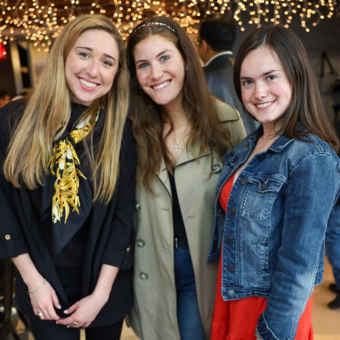 Three young women smiling at a social gathering, with string lights in the background.