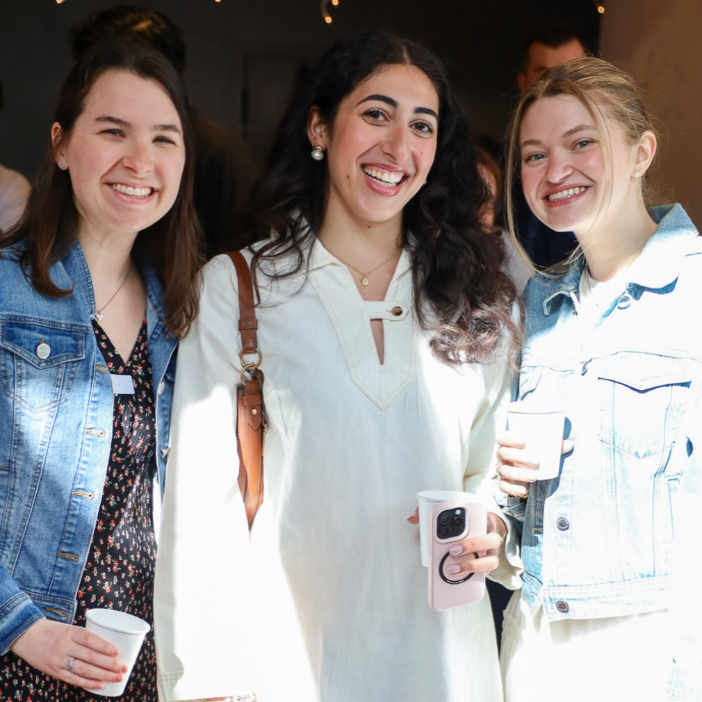 Three women smiling at a social event, holding cups and a phone, dressed casually