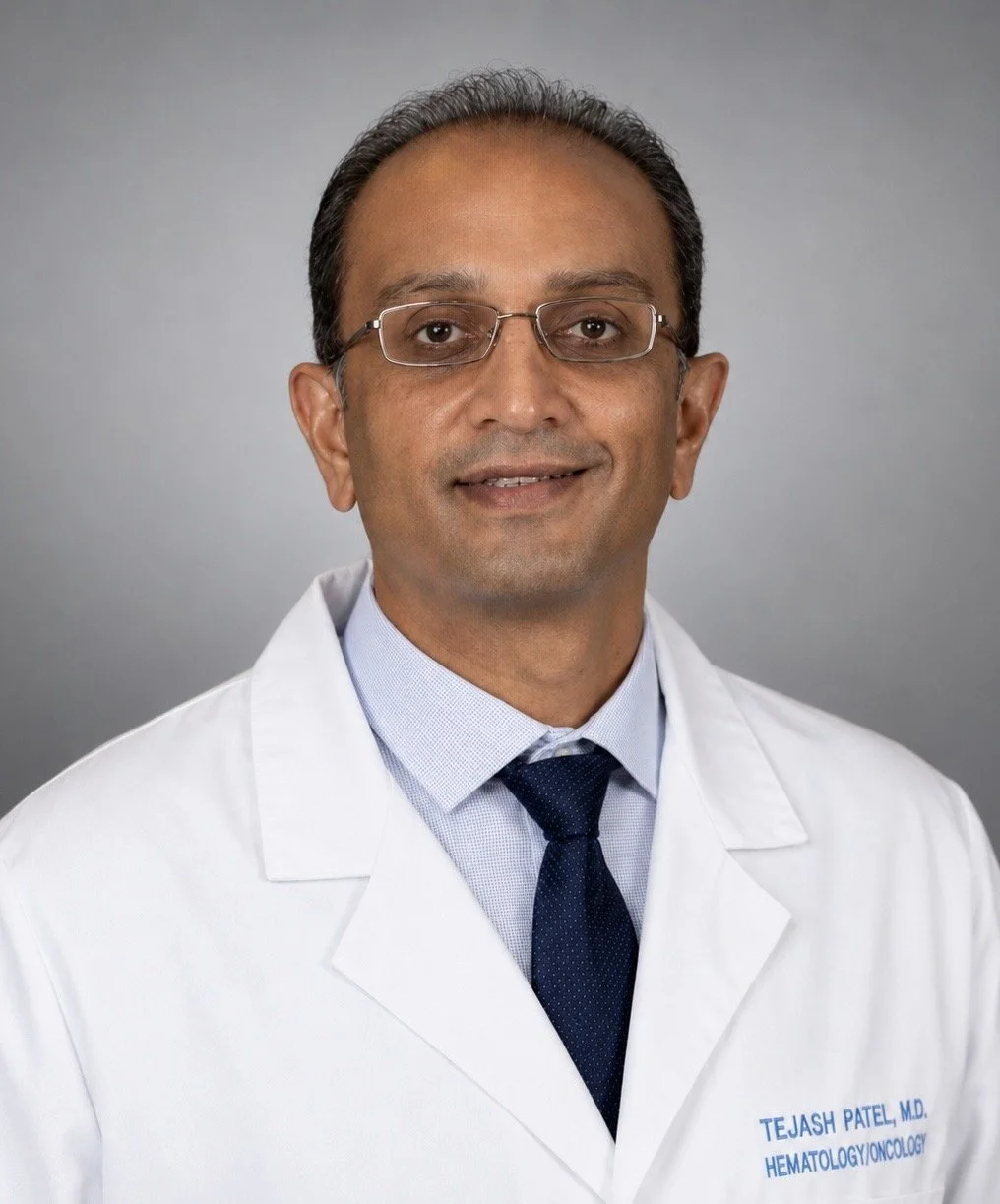 Professional portrait of Dr. Tejash Patel in a white medical coat with a blue shirt and dark blue tie, set against a plain gray background.