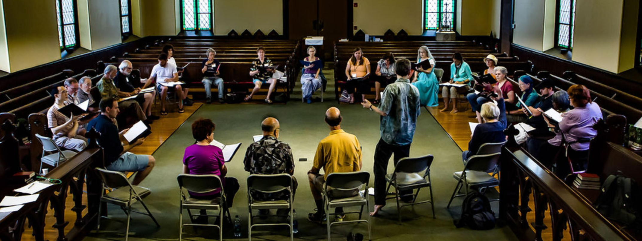 Group of people seated in a large circle, participating in a discussion or workshop, in a spacious room with wood floors and stained glass windows.