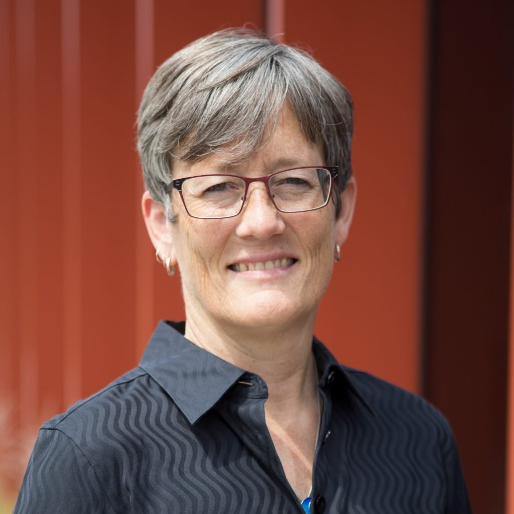 A portrait of a middle-aged woman with short gray hair, wearing glasses, earrings, and a dark collared shirt, smiling in front of a red background.