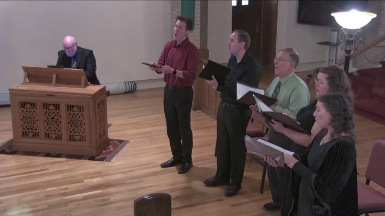 A group of five people performing music in a room with wooden floors. One person is seated at a keyboard, while the others are standing with songbooks.