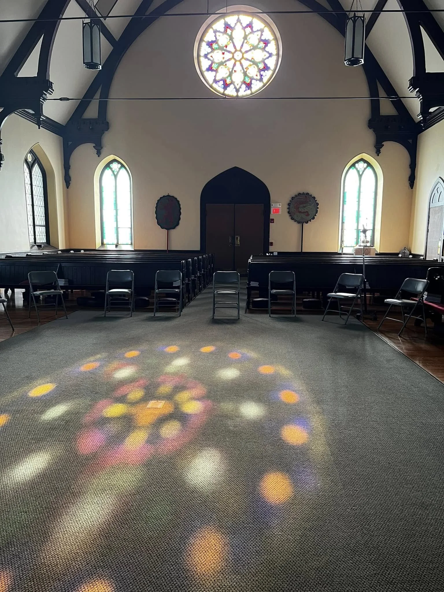 Interior of a church or chapel with stained glass windows, wooden pews, a carpeted floor, and a large circular stained glass window at the front.
