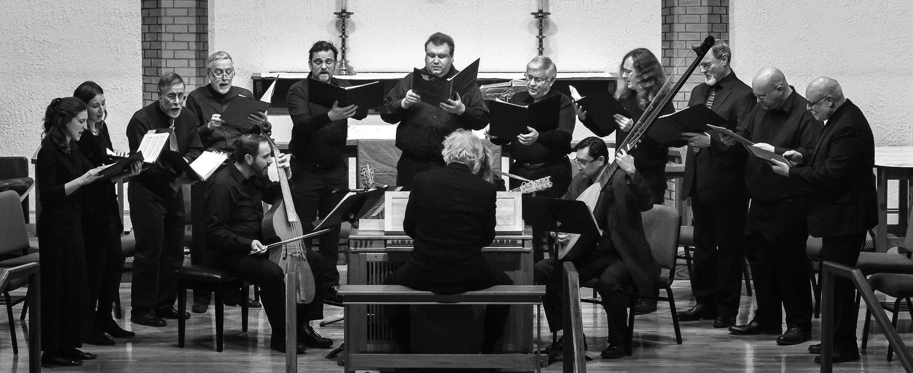 A choir and musicians performing in a church or concert hall. The choir members are standing and holding sheet music, with some playing string instruments, while the conductor directs the performance.
