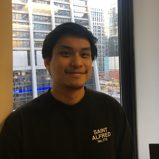 A young man standing indoors near a window with a cityscape view, wearing a black sweatshirt with white text that reads 'SAINT ALFRED No.773'.