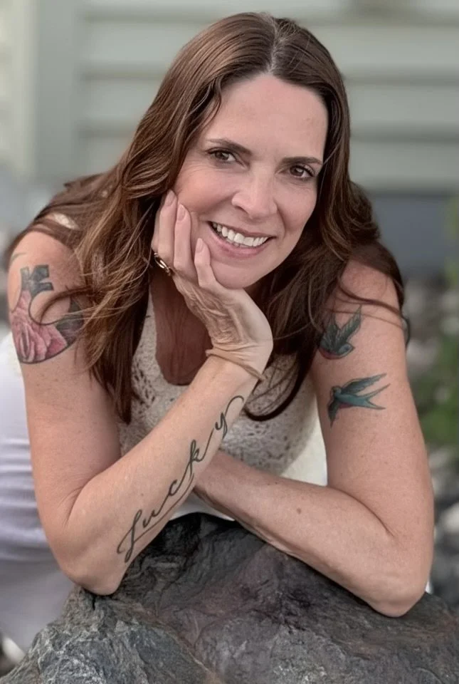A woman with brown hair smiling, resting her head on her hand, wearing a sleeveless top. She has tattoos on her arms, including birds and a script tattoo on her forearm. She is outdoors, leaning on a large rock.