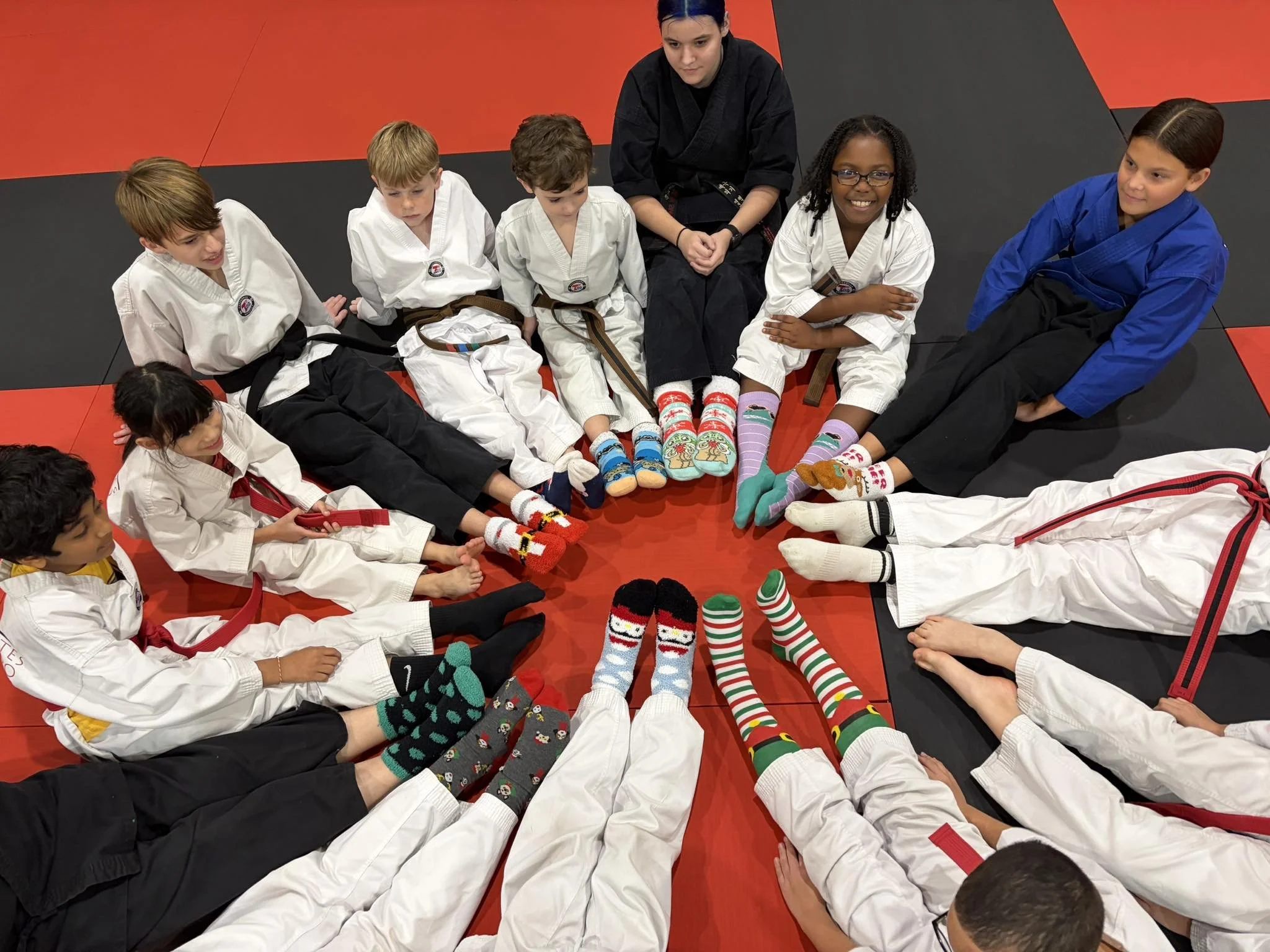 Children dressed in martial arts uniforms sitting on a red and black mat with their legs extended forward, wearing colorful festive socks, in a martial arts dojo.