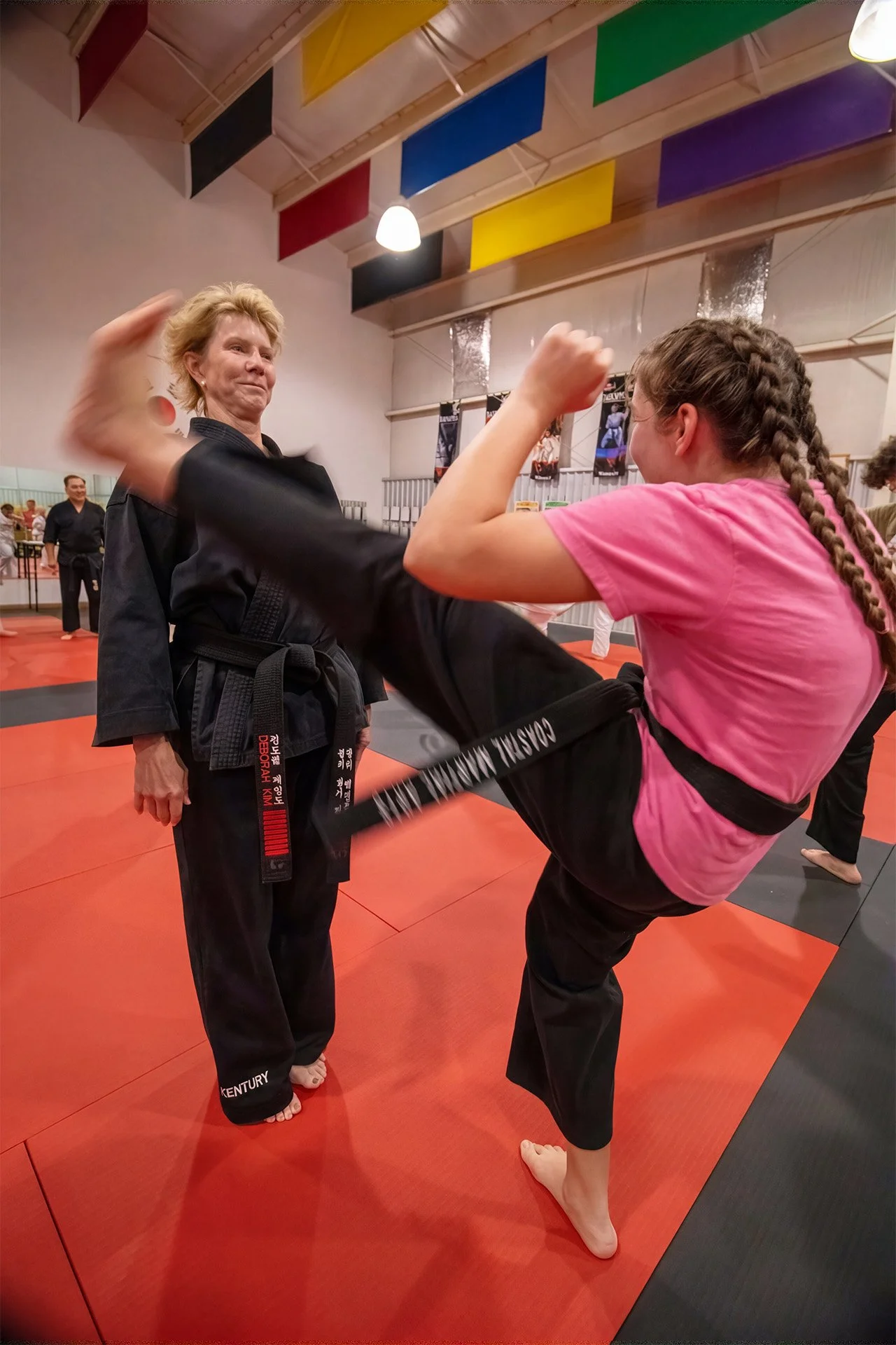 A young girl practicing a high kick in a martial arts training room, supervised by an older female instructor in a black uniform. Other students are visible in the background, and colorful banners hang from the ceiling.