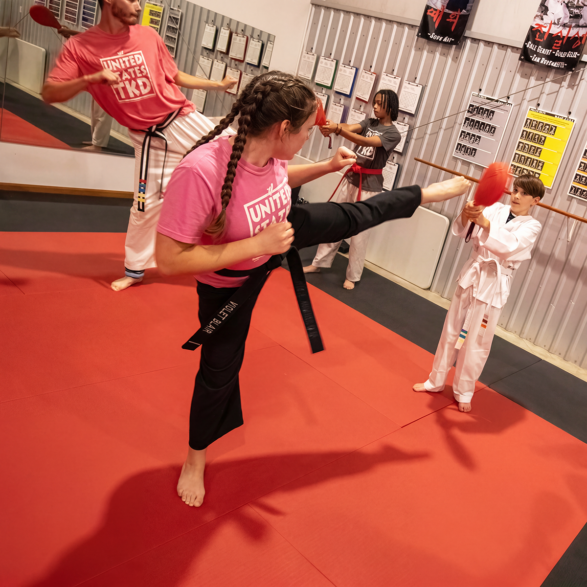 Kids practicing martial arts in a dojo, with girl performing a high kick and other children holding targets and sticks.