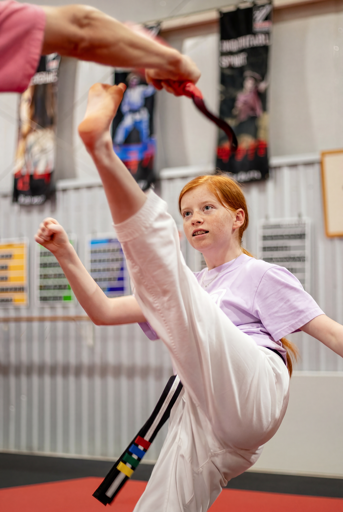 A young girl with red hair is practicing martial arts in a dojo, executing a high kick while holding a black stick with colored segments.