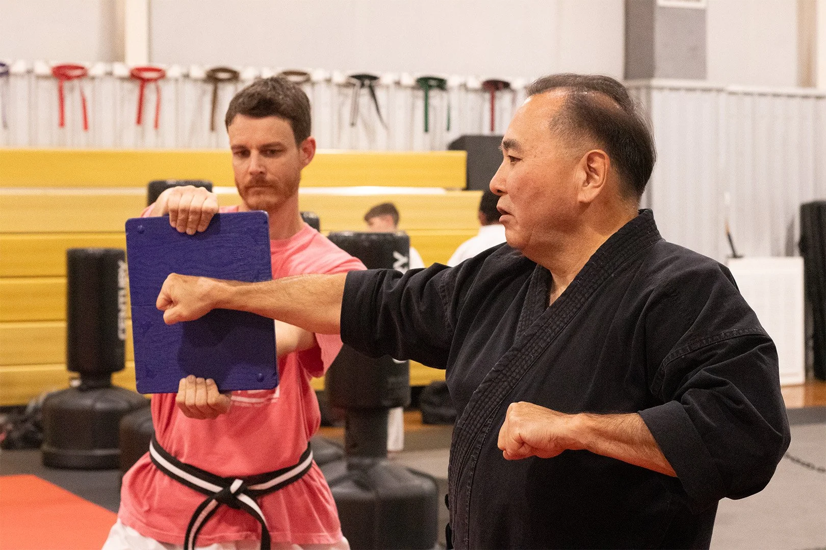 Martial arts instructor demonstrating a punch technique with a training pad with a student in a gym with punching pads and gym equipment.