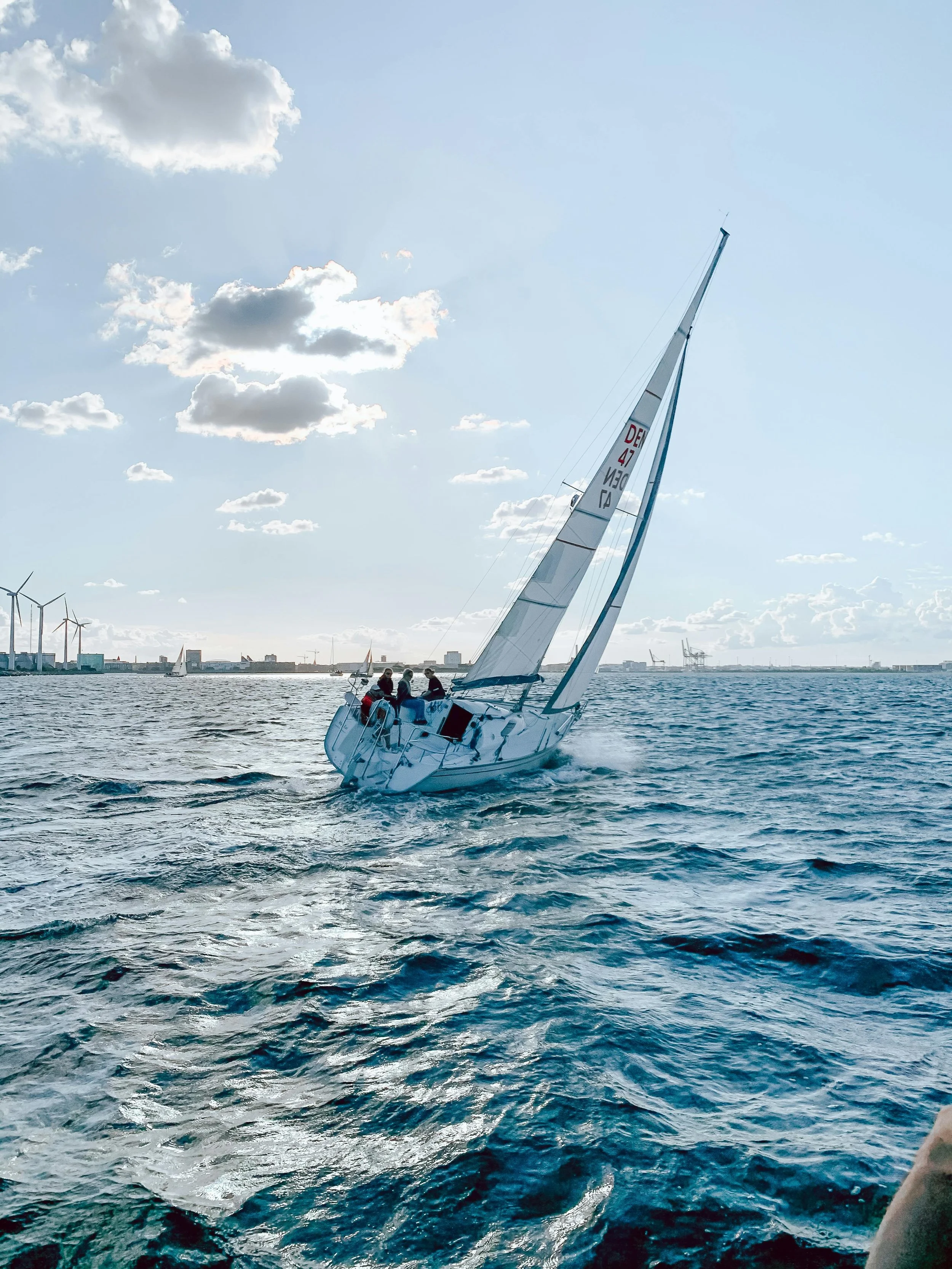 A sailboat with three people on board sailing on the water under a partly cloudy sky, with wind turbines visible in the background.
