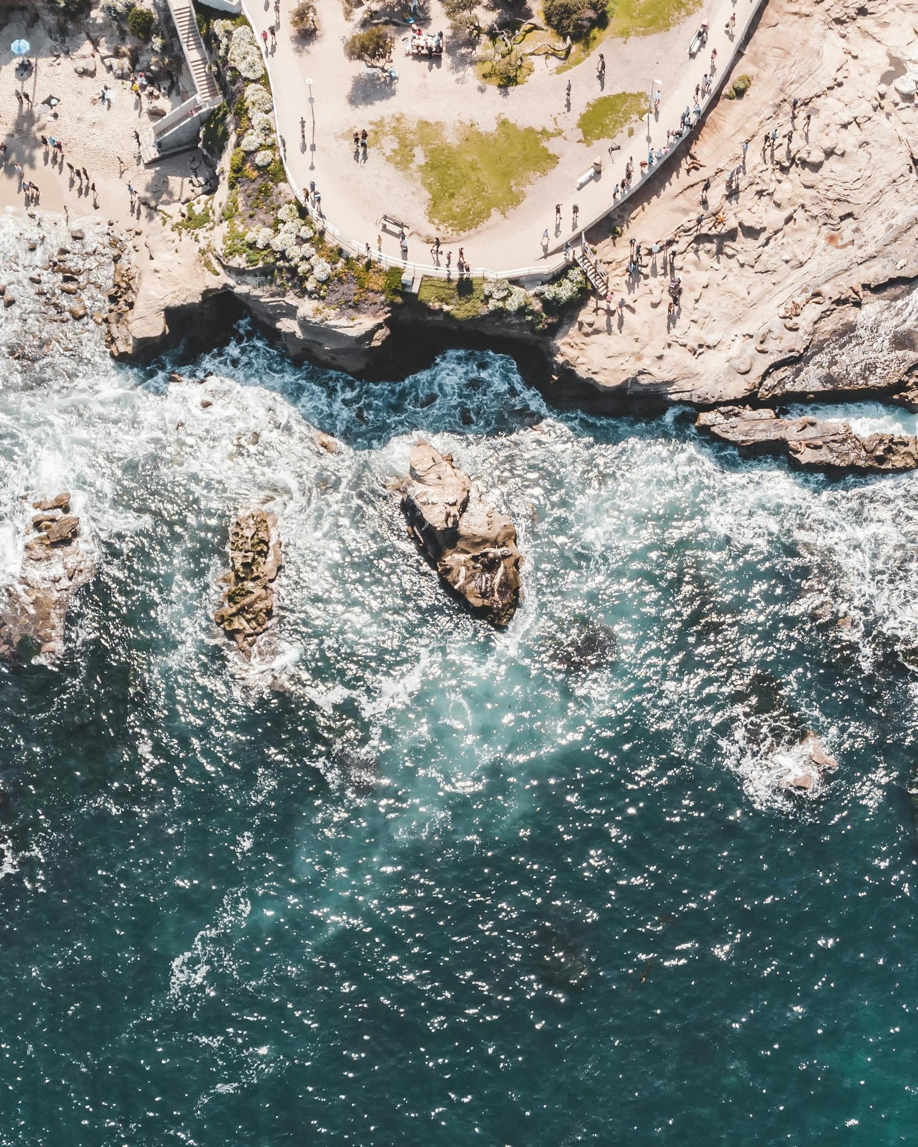 An aerial view of a park with a small crowd of people walking along pathways, surrounded by rocky terrain and ocean waves crashing against rocks below.