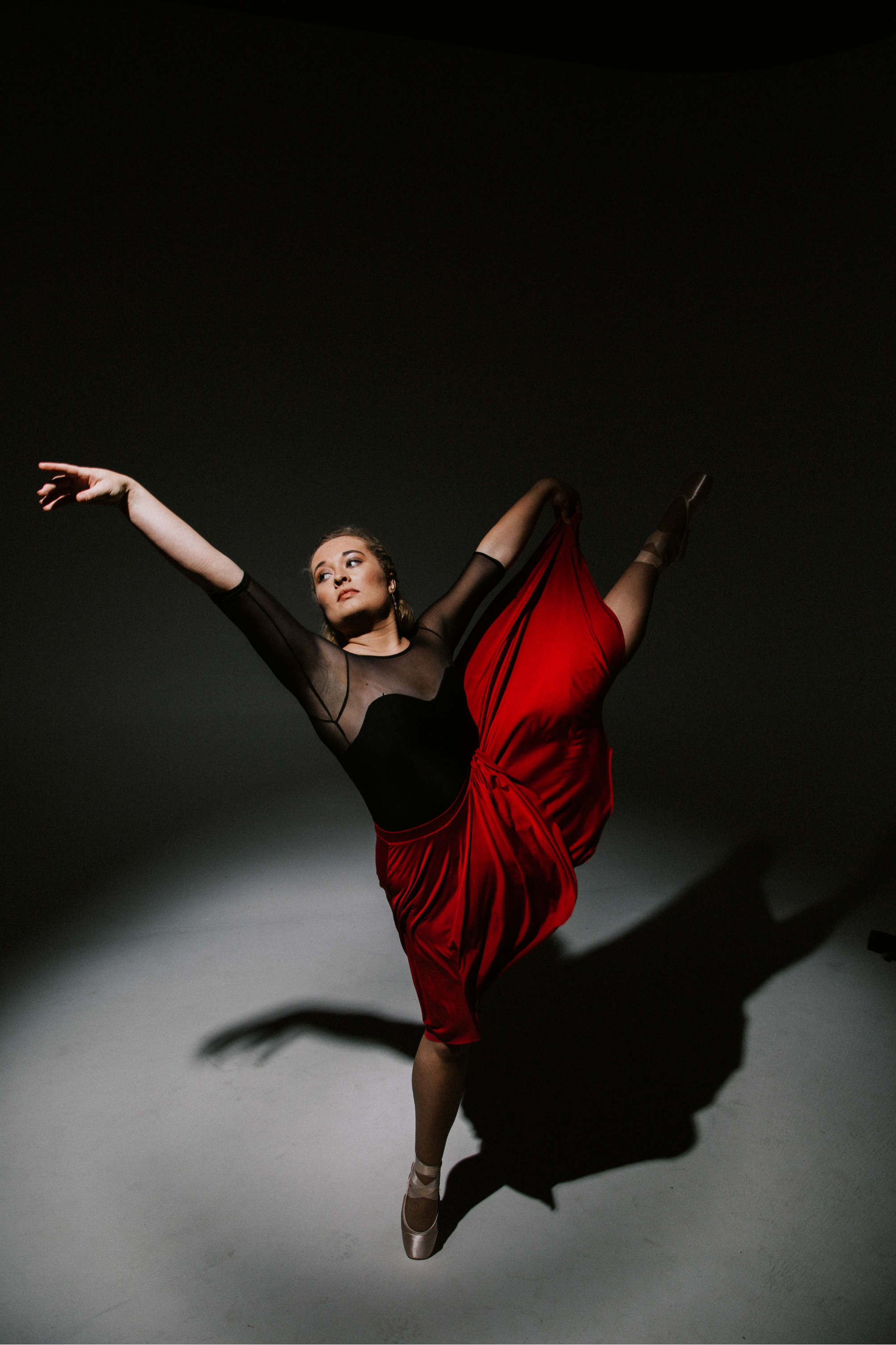 A ballerina in a black long-sleeve leotard and red dance skirt performing an arabesque pose, standing on pointe, against a dark background with a shadow cast on the floor.