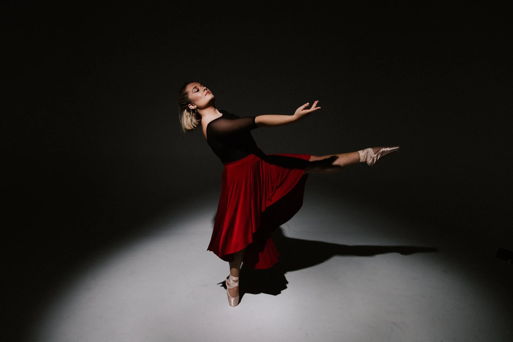 A ballet dancer in a black and red costume posing on the tips of her toes against a dark background.