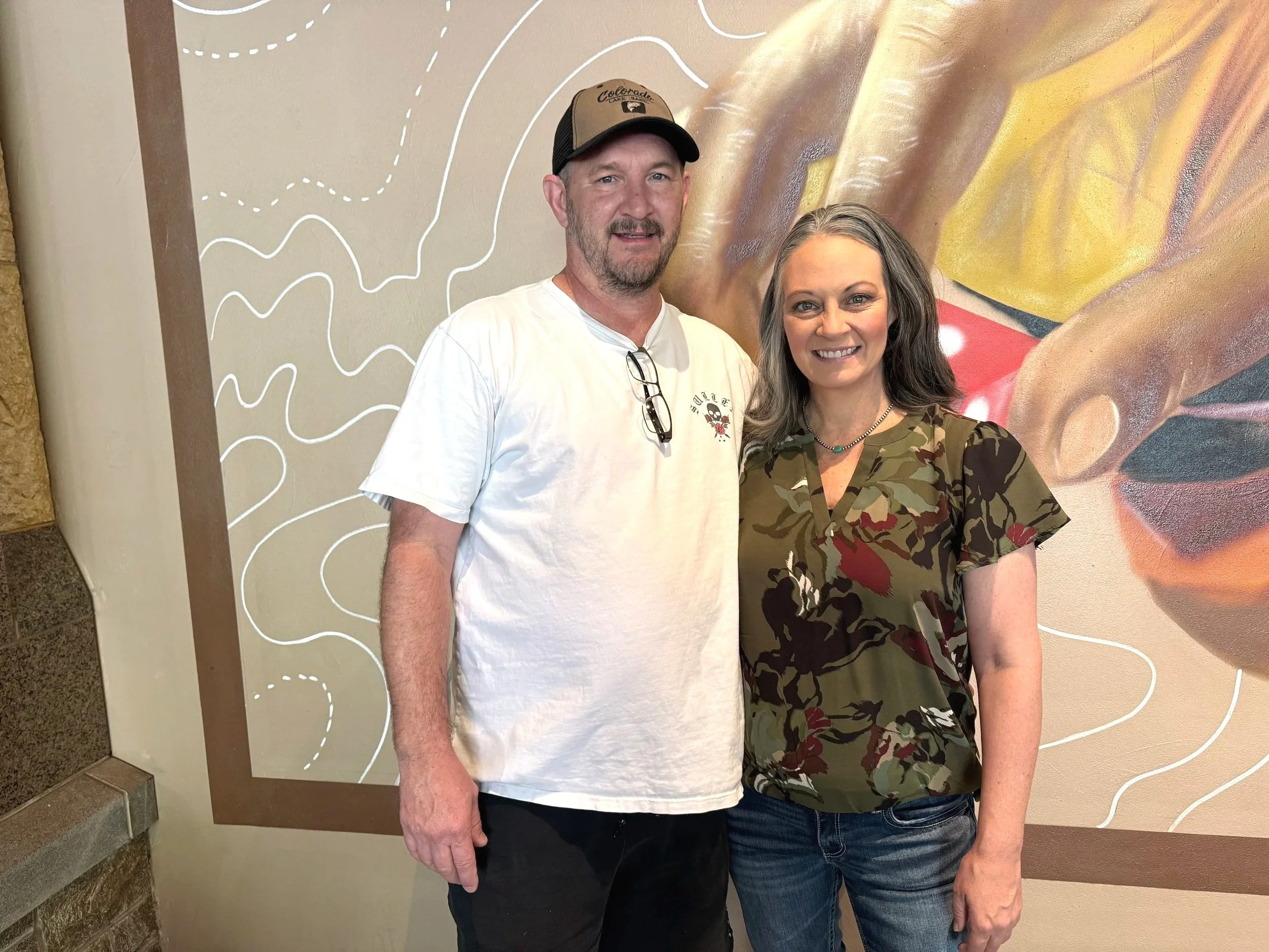 A man and a woman standing together indoors in front of a colorful wall with abstract art. The man is wearing a white t-shirt and a baseball cap, while the woman is dressed in a floral camouflage patterned shirt and has gray hair.