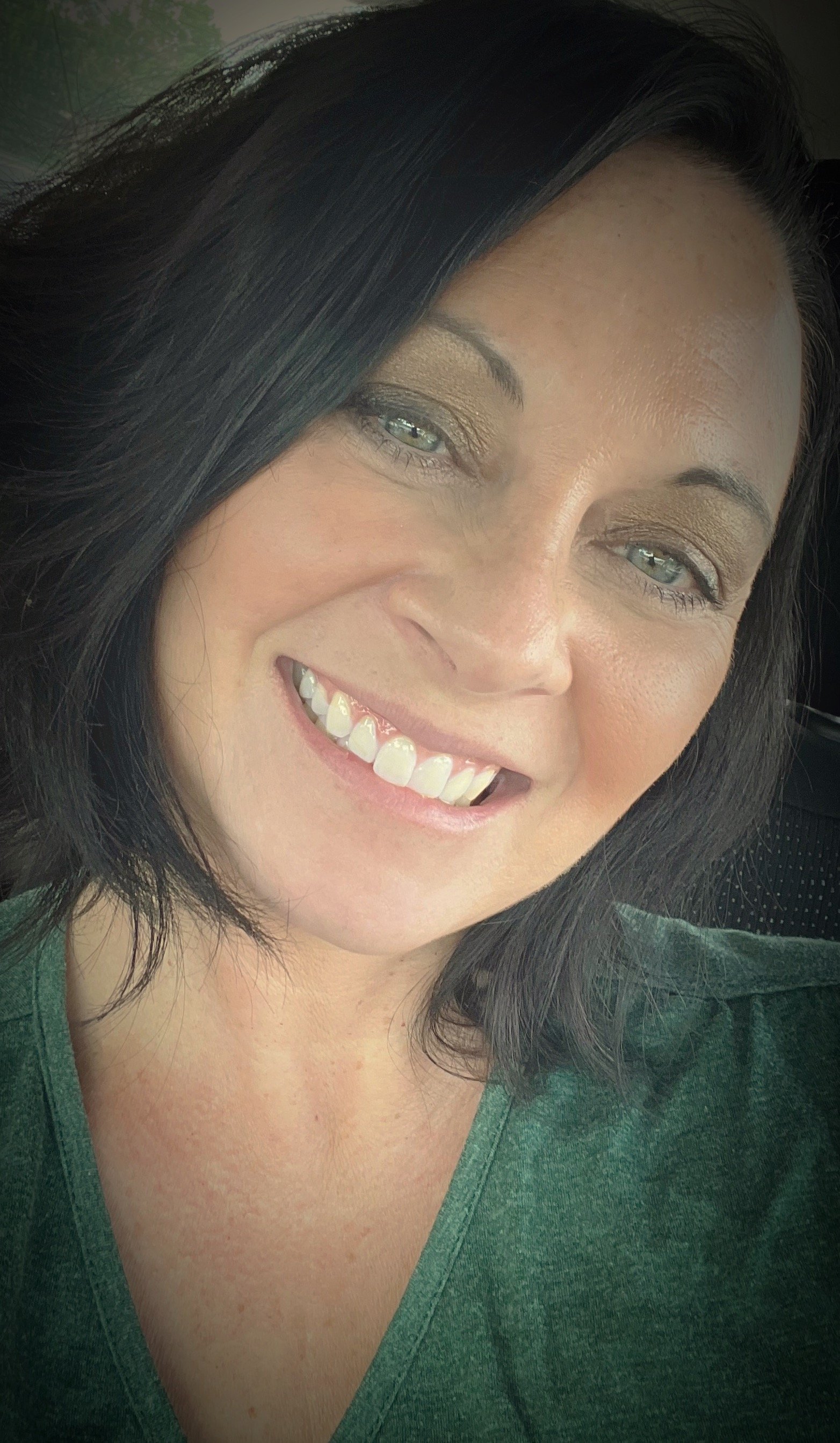 Close-up of a smiling woman with dark hair and light skin, showing her teeth and wearing a green top.