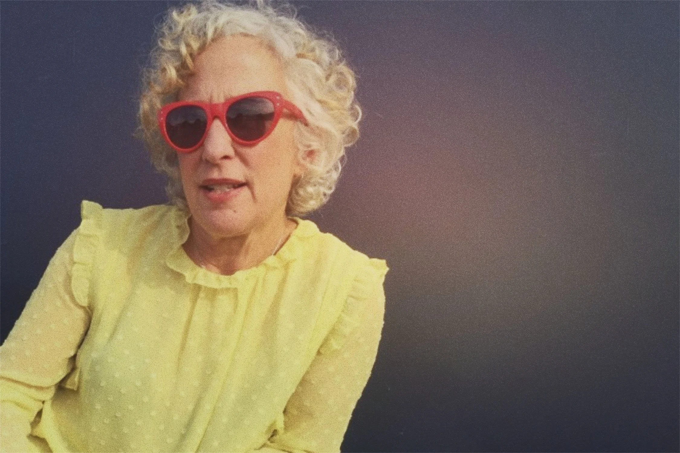 A woman with curly white hair wearing red sunglasses and a yellow blouse, against a plain dark background.