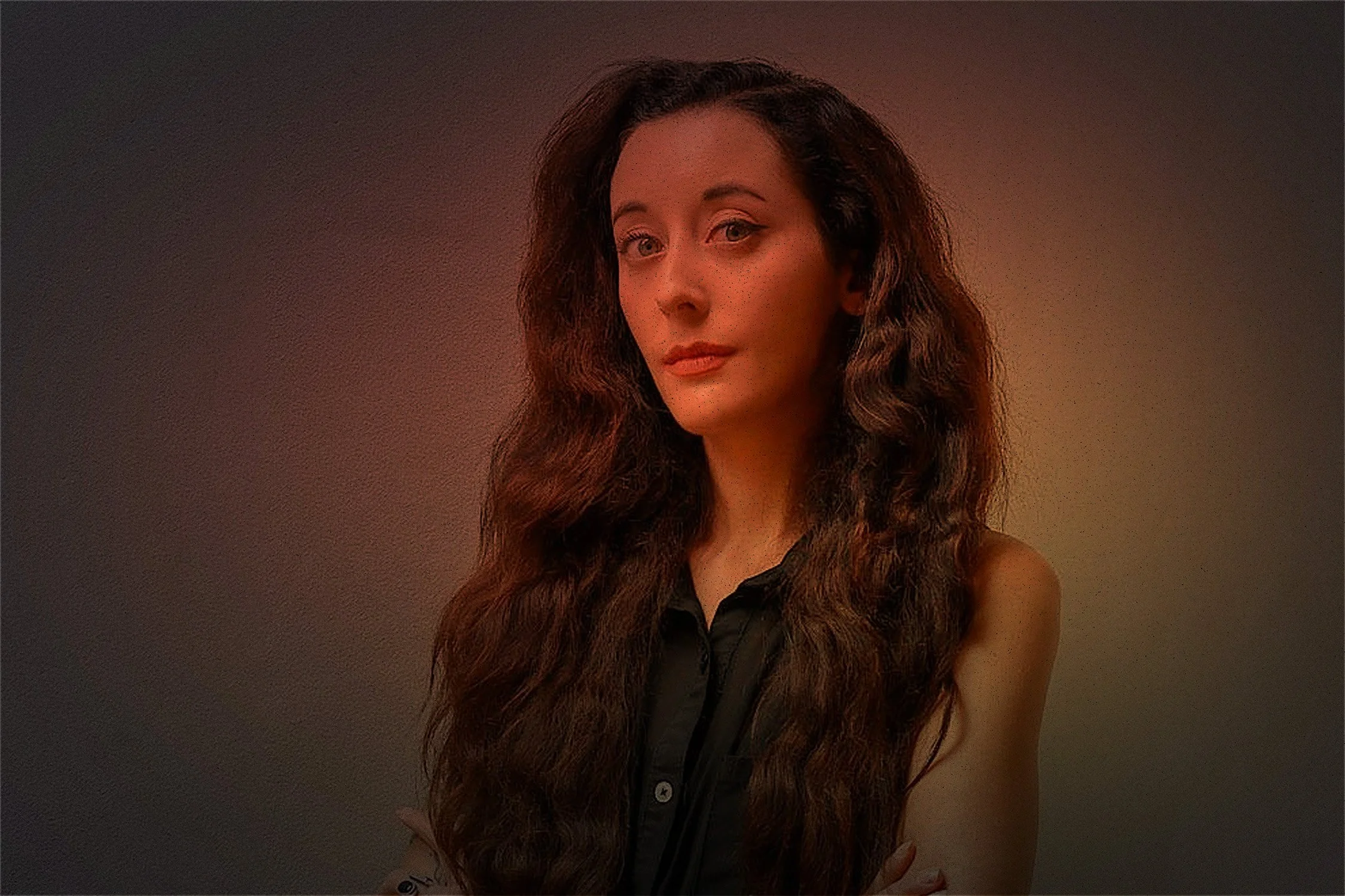 A woman with long, wavy brown hair, wearing a sleeveless black shirt, standing against a plain wall.