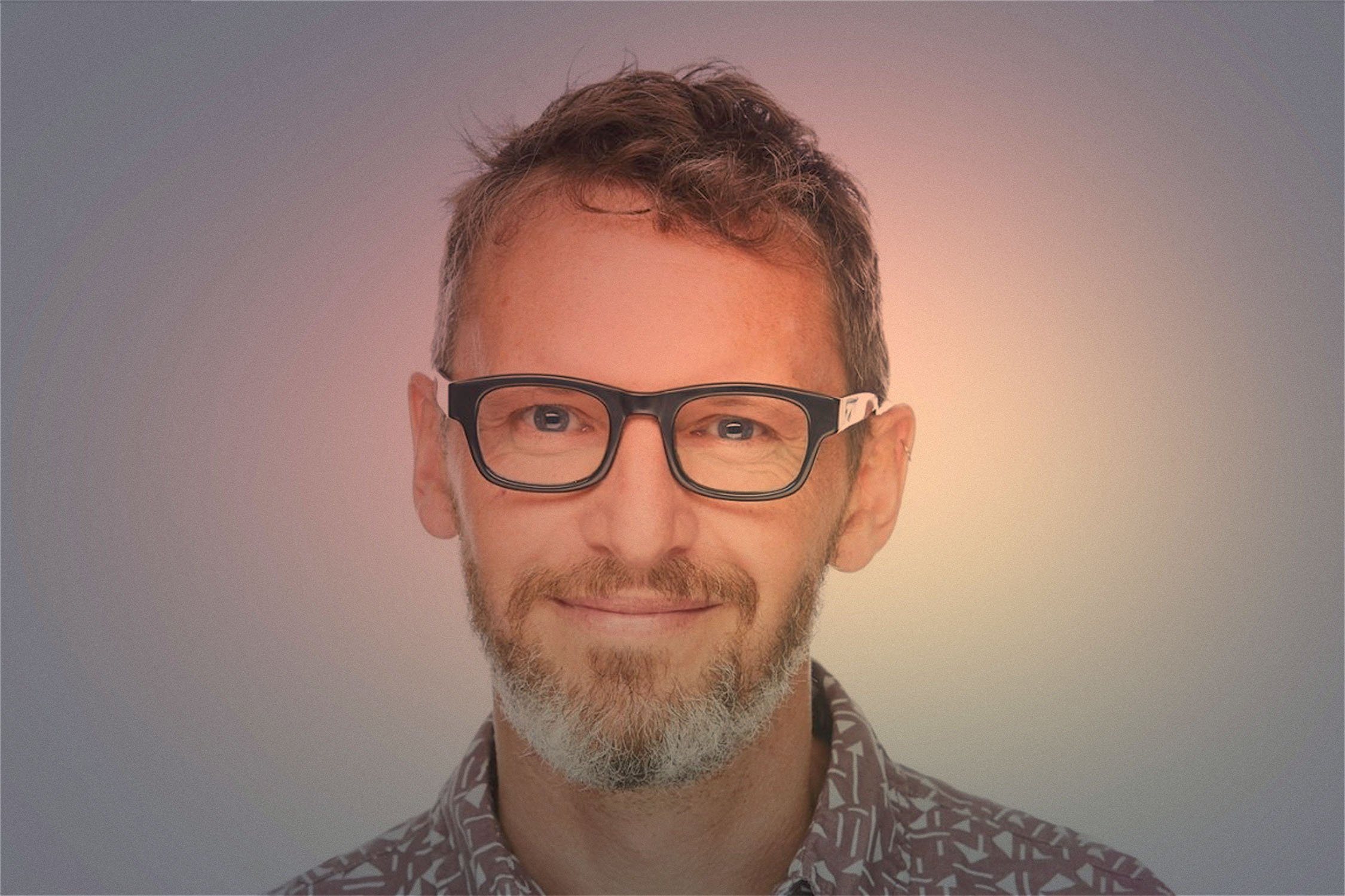 A man with light skin, short curly brown hair, a beard, and glasses with black frames, smiling and wearing a patterned shirt