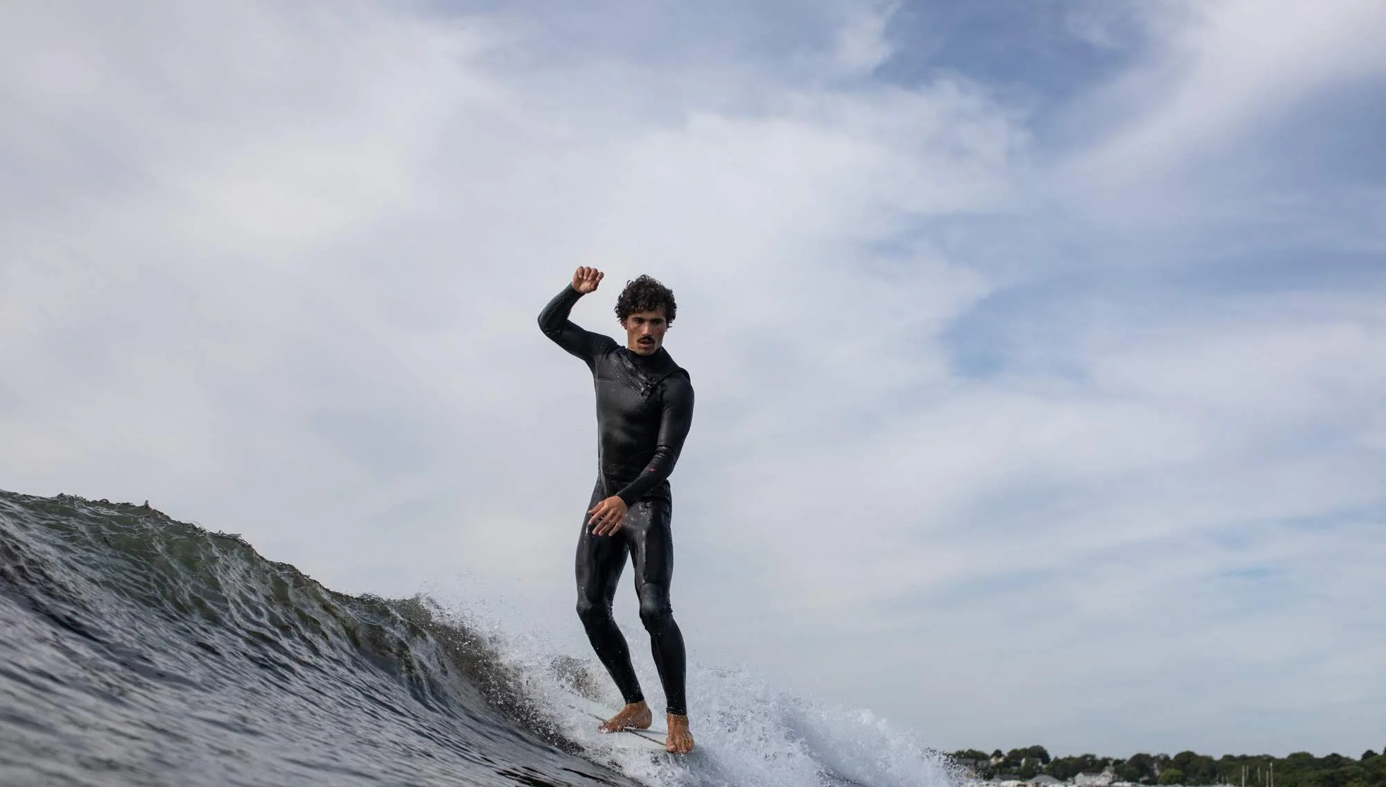 A man surfing on a wave in the ocean, wearing a black wetsuit, with a cloudy sky overhead.