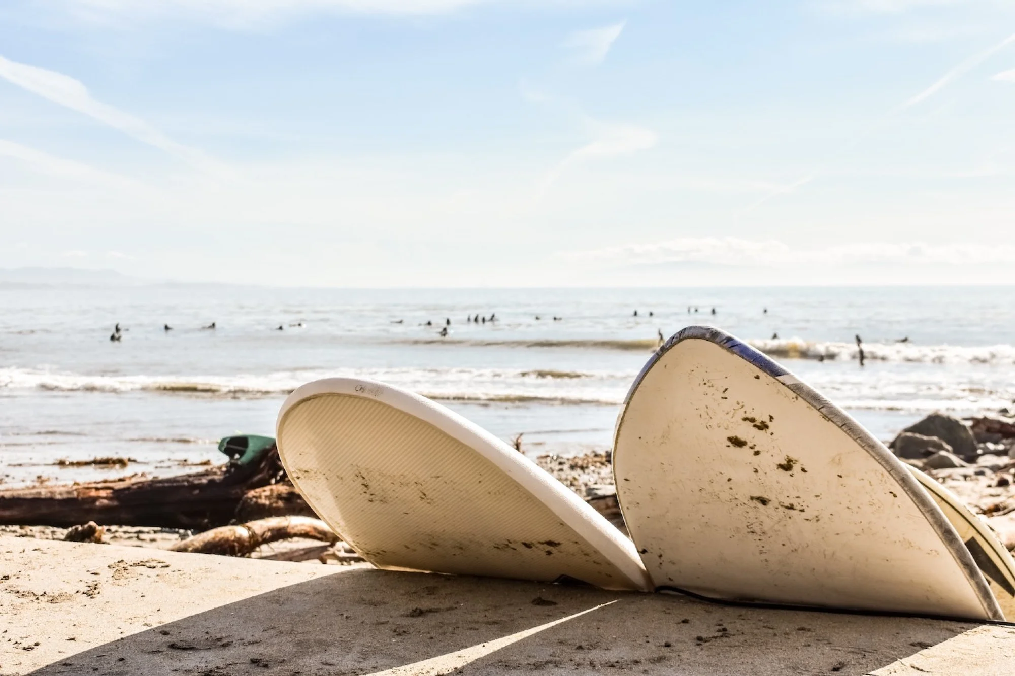 Two surfboards resting on the sandy beach near driftwood, with the ocean and a clear blue sky with some clouds in the background. In the distance, people are swimming and surfing in the water.