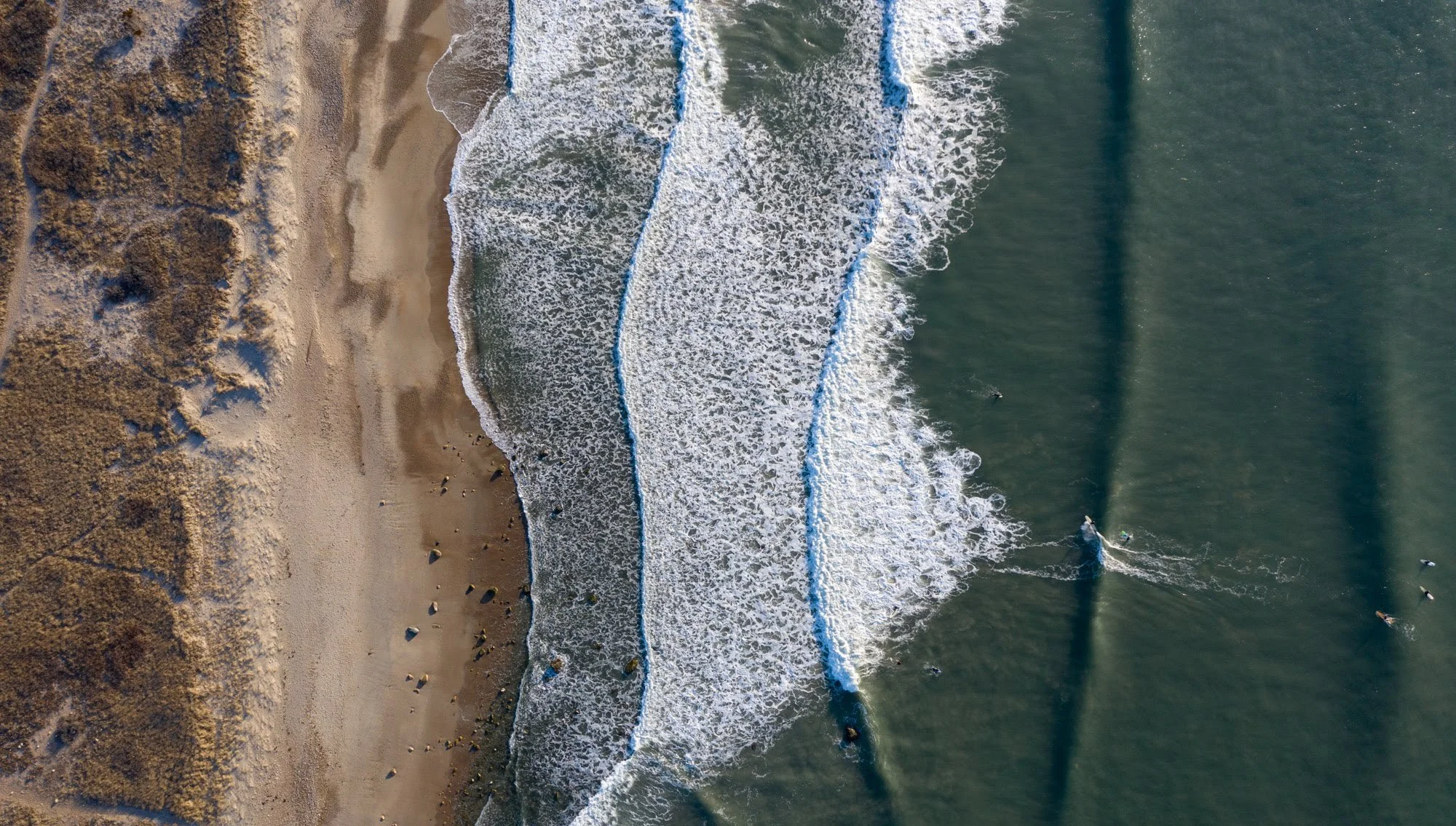 Aerial view of a beach with sandy shore, waves breaking, and surfers in the water.